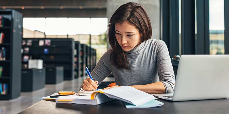 Teacher preparing the next class with a book and taking notes. She wears a grey turtleneck jumper.