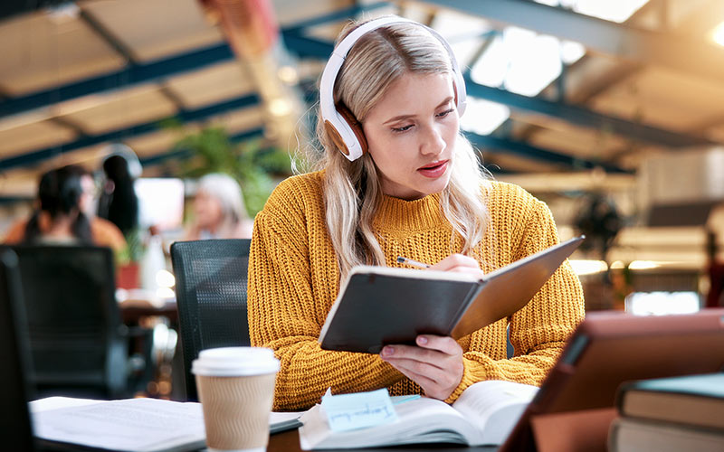 Learner wearing headphones reviewing study materials and taking notes, with a tablet and books on the table.
